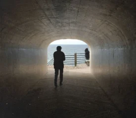 Silhouetted person walking through a dark tunnel toward a bright ocean view, with another figure standing near the railing outside