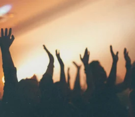 Silhouetted crowd with raised hands in a dimly lit room, illuminated by warm stage lighting