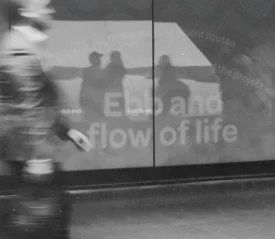 A blurred figure walks past a reflective wall displaying the words “Ebb and flow of life,” with silhouettes visible in the reflection