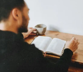 A person sits at a wooden table reading an open Bible with a mug beside them and a smartphone resting nearby