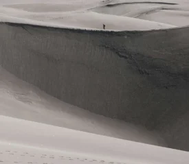 Person walking along the crest of a massive sand dune in a vast, windswept desert landscape