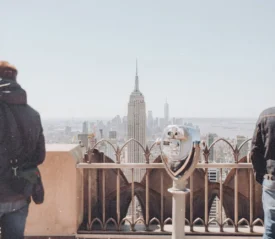 Two people standing at an observation deck overlooking the New York City skyline, with the Empire State Building centered in the view and a coin‑operated viewer in the foreground