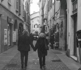 Two people walking hand‑in‑hand down a narrow cobblestone street lined with shops and tall historic buildings