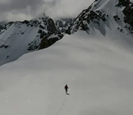A lone person walks across a wide snowy slope surrounded by towering, rugged mountains and low-hanging clouds