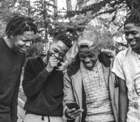 Four people stand outdoors closely together, looking at a smartphone one person is holding