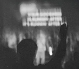 Silhouetted person raising one hand in front of a brightly lit stage with indistinct figures and text in the background
