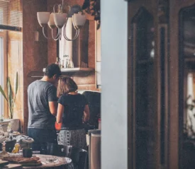 A couple standing together in a cozy kitchen preparing food at the counter, surrounded by warm lighting and houseplants