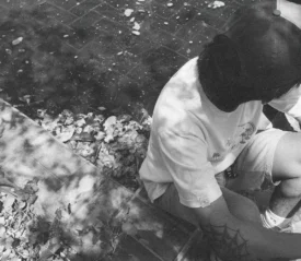 Black‑and‑white photo of a person sitting on outdoor steps in dappled sunlight, with fallen leaves scattered around