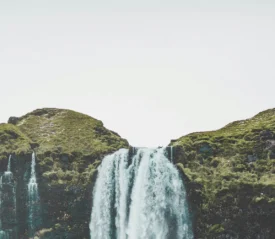 Wide waterfall cascading over a grassy cliff into a rocky valley under a bright, overcast sky