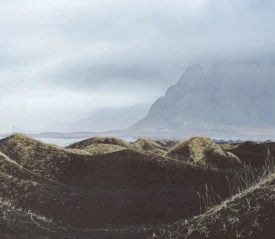 Wind-shaped black sand dunes under a cloudy sky with a mist-covered mountain in the distance