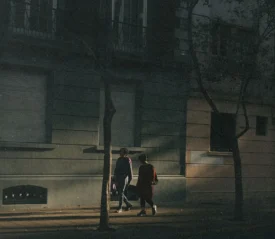 Two people walk side by side down a dimly lit urban street, illuminated by a beam of late‑day sunlight falling across the sidewalk