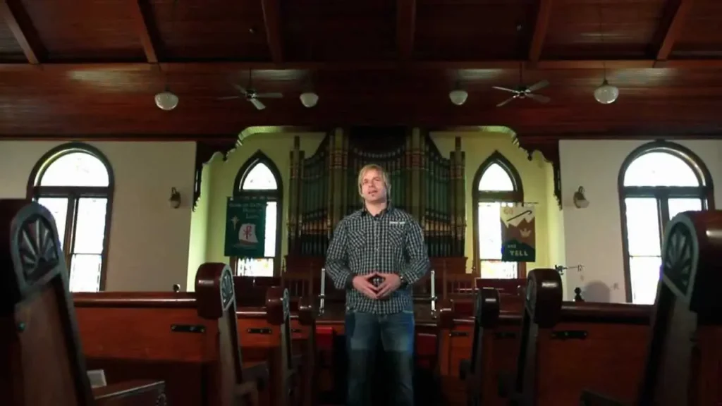 Man standing in a church aisle, hands clasped, with organ pipes and wooden ceiling in the background.