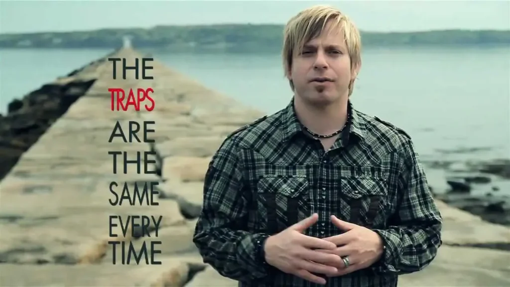 Man in a plaid shirt stands on a rocky pier by the water, hands clasped, with the line “THE TRAPS ARE THE SAME EVERY TIME” overlaid.