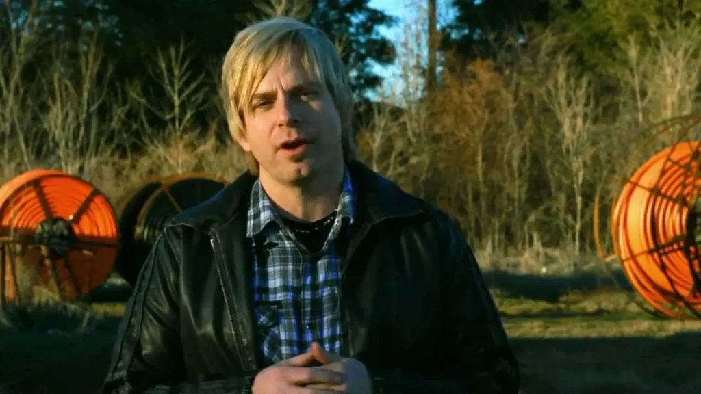 Blond man in a leather jacket speaks to the camera outdoors, with orange cable reels in the background.