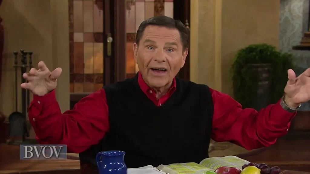 Televangelist in a studio, wearing a red shirt and black vest, speaking with animated hand gestures over an open Bible on a table with fruit and a blue mug nearby