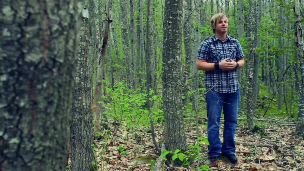 Man in a plaid shirt and blue jeans stands among trees in a forest, hands clasped, looking off to the right