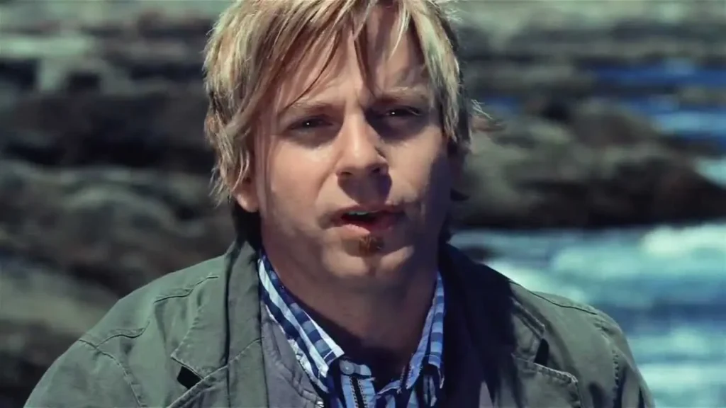 Man with blond hair wearing a green jacket and checkered shirt by the rocky shoreline, cigarette in mouth, looking toward camera.