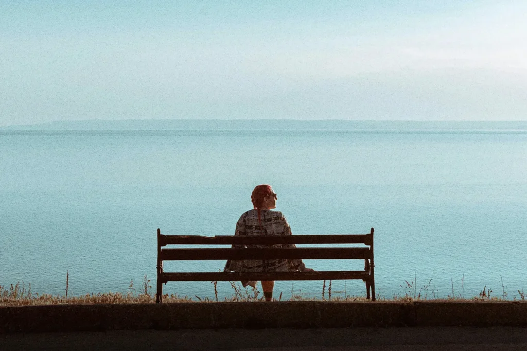 Person sitting alone on a bench overlooking calm blue water, creating a quiet moment that reflects self-care, mental health, patience, and realignment.