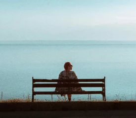 Person sitting alone on a bench overlooking calm blue water, creating a quiet moment that reflects self-care, mental health, patience, and realignment.