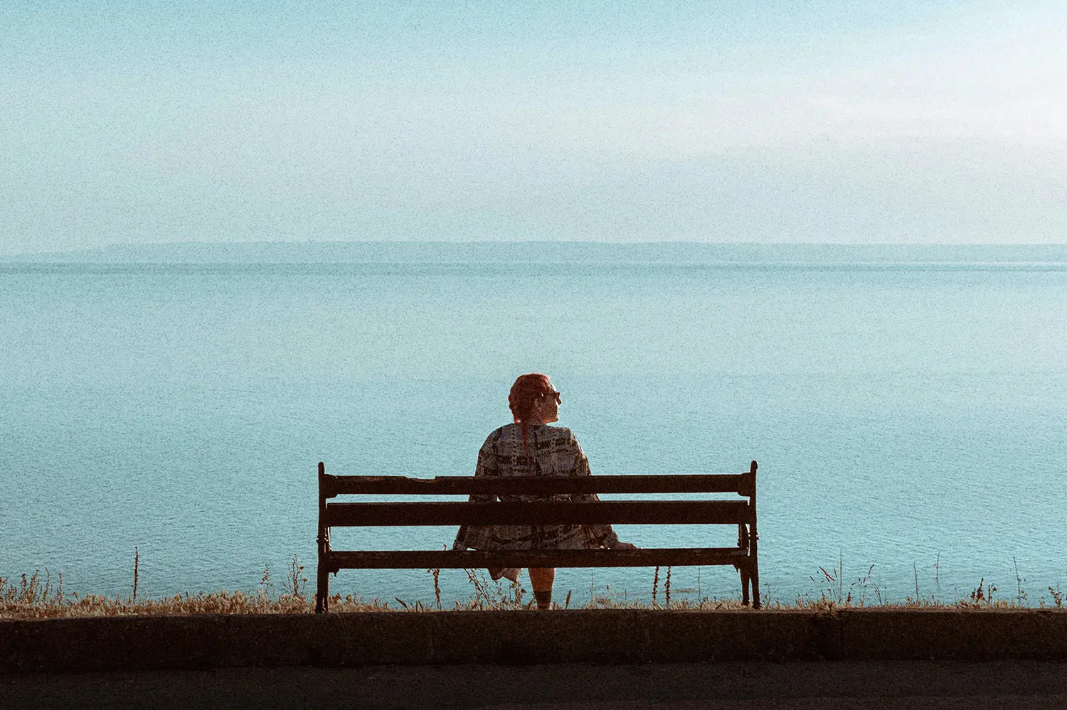 Person sitting alone on a bench overlooking calm blue water, creating a quiet moment that reflects self-care, mental health, patience, and realignment.