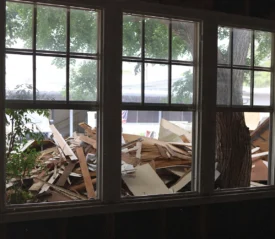 View through interior windows showing piles of storm debris, broken wood, and fallen materials outside, illustrating disaster relief needs after severe weather impacts.