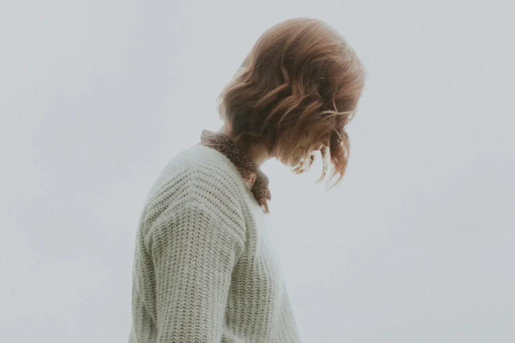 Person with shoulder-length light brown hair wearing a cream knit sweater and a patterned scarf, looking downward.