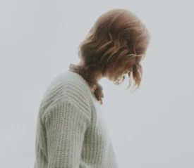 Person with shoulder-length light brown hair wearing a cream knit sweater and a patterned scarf, looking downward.