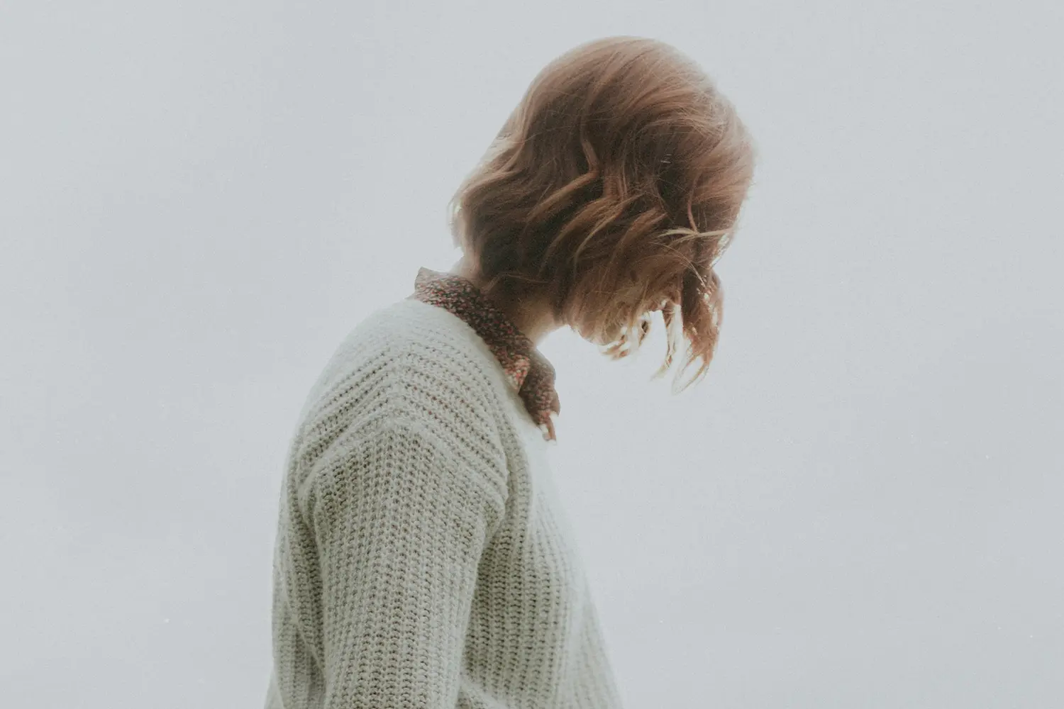 Person with shoulder-length light brown hair wearing a cream knit sweater and a patterned scarf, looking downward.