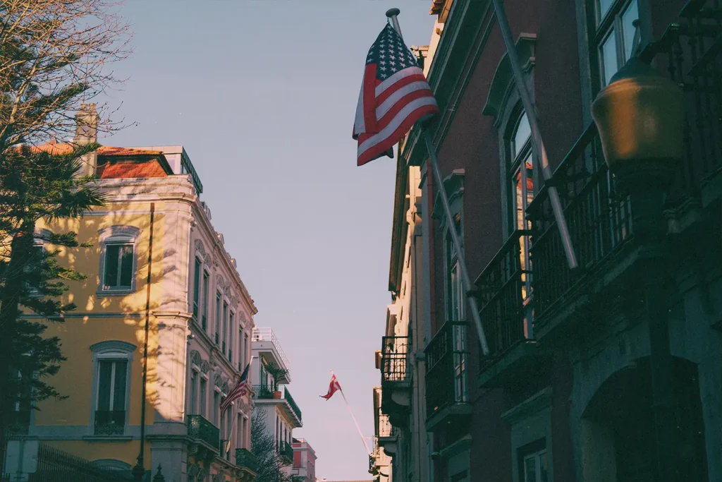 Street view with colorful buildings and prominent waving American flag overhead; a second flag in the distance.