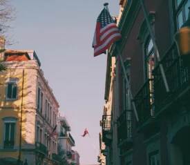 Street view with colorful buildings and prominent waving American flag overhead; a second flag in the distance.