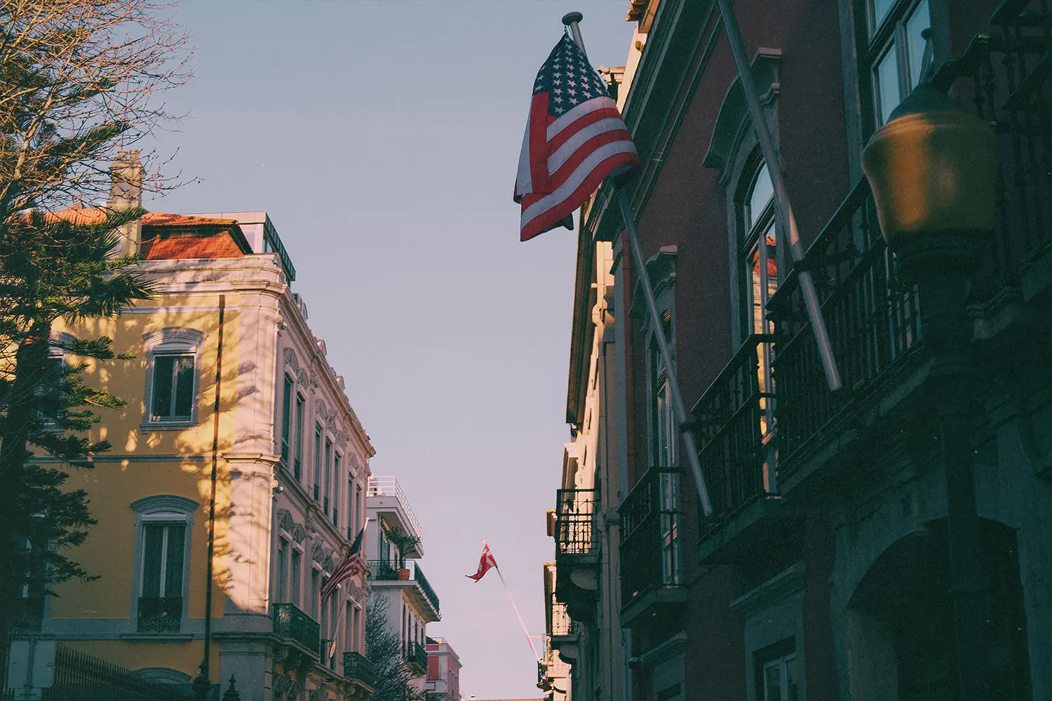 Street view with colorful buildings and prominent waving American flag overhead; a second flag in the distance.