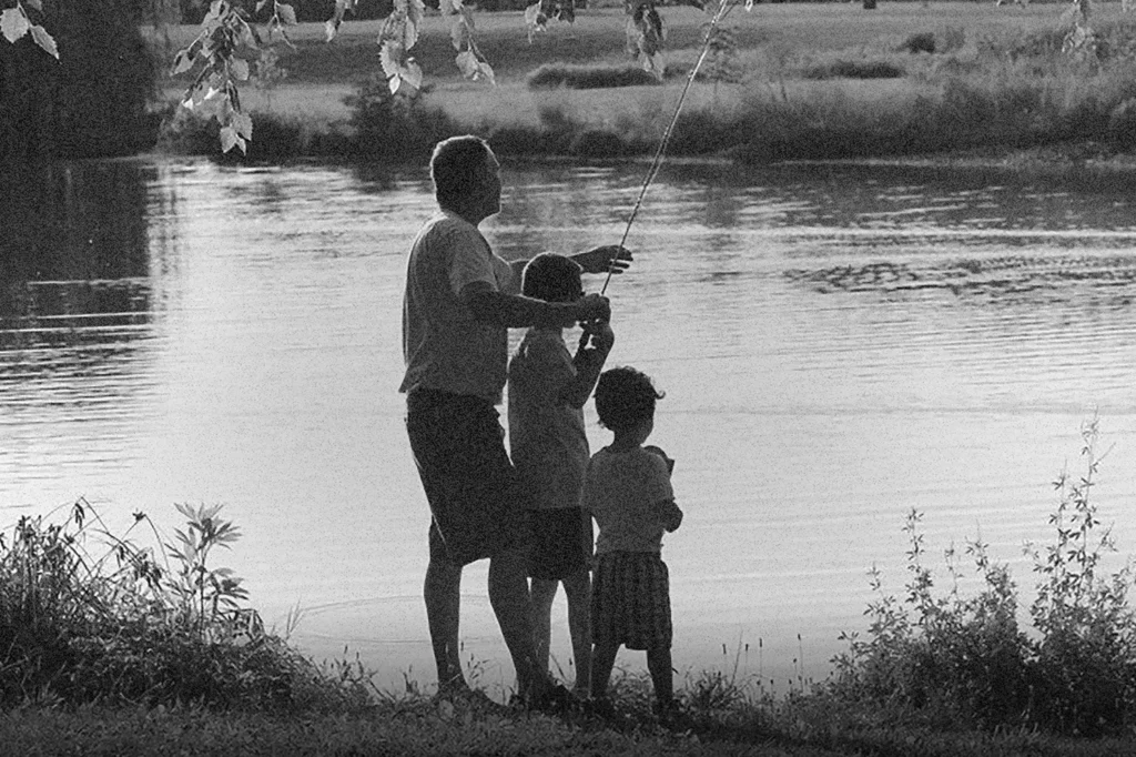 Three children stand at the edge of a lake, one holding a fishing rod while the others watch by the water’s edge.