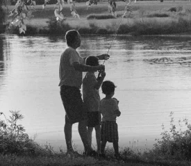 Three children stand at the edge of a lake, one holding a fishing rod while the others watch by the water’s edge.