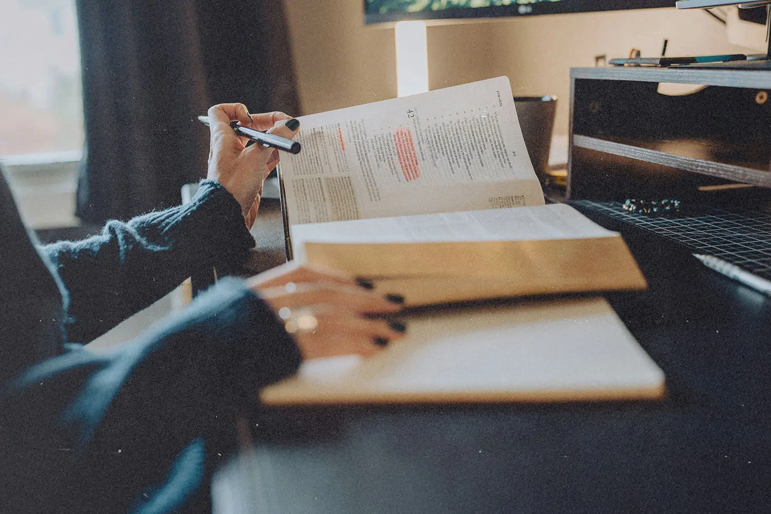 A person studies at a desk with an open Bible, highlighting passages while writing notes in a notebook