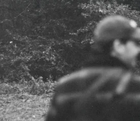 Man wearing a hat, standing in a wooded clearing with dense trees in the background