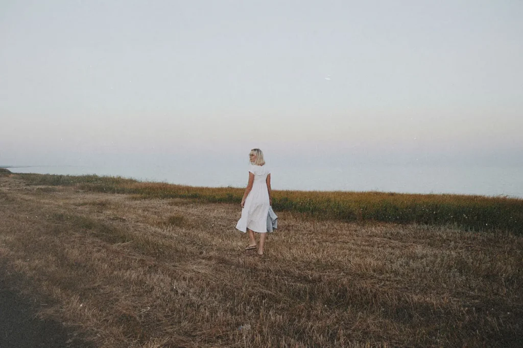 A woman in a white dress walks through a dry, open field near the shoreline under a pale evening sky
