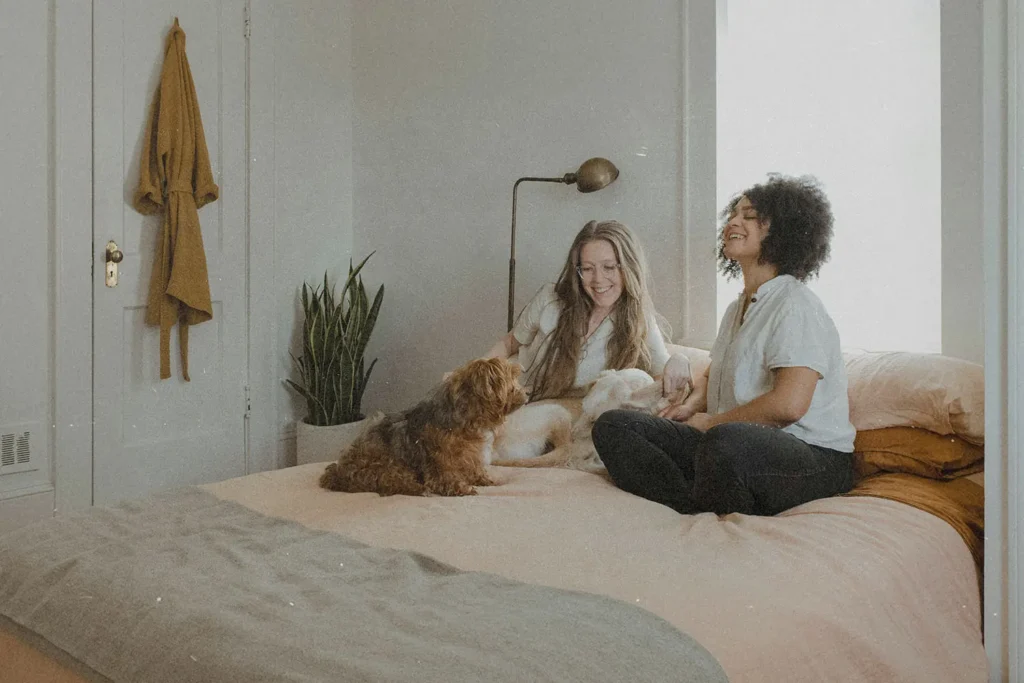 Two people sitting on a bed with two small dogs in a softly lit bedroom