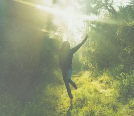 A person stands on a sunlit forest path with arms raised, surrounded by tall trees and bright rays of light streaming through the foliage