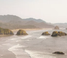 Ocean waves roll toward a sandy shoreline with large rocky sea stacks and distant mountains under a hazy sky