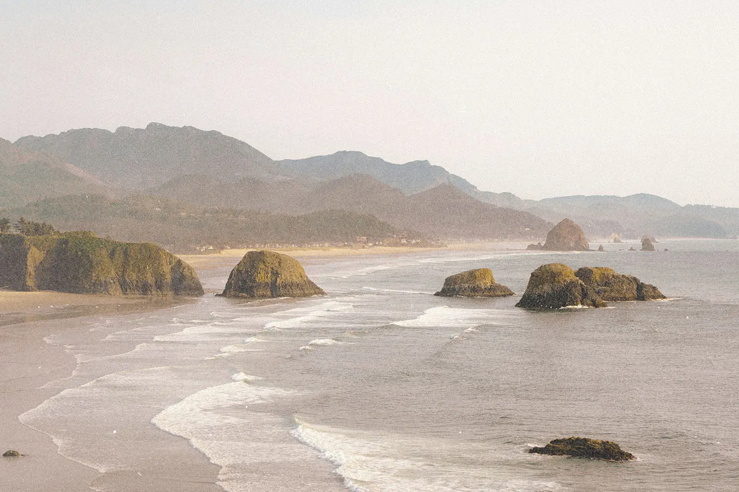 Ocean waves roll toward a sandy shoreline with large rocky sea stacks and distant mountains under a hazy sky