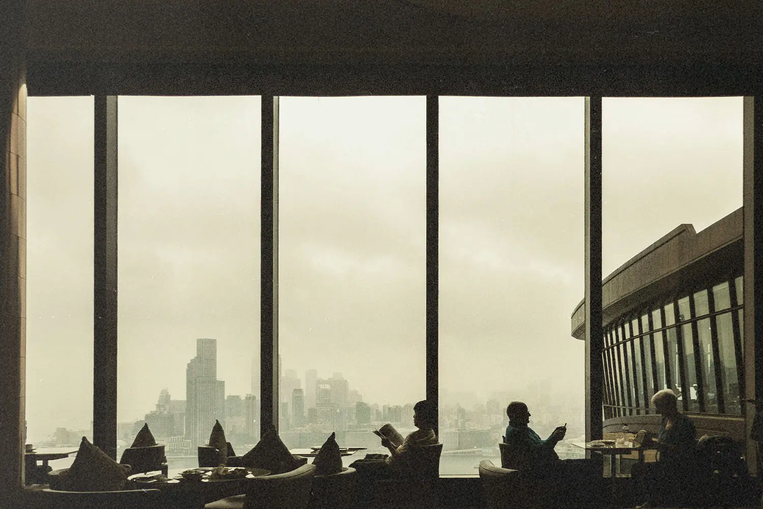 People sit at tables inside a restaurant with tall windows overlooking a cloudy city skyline