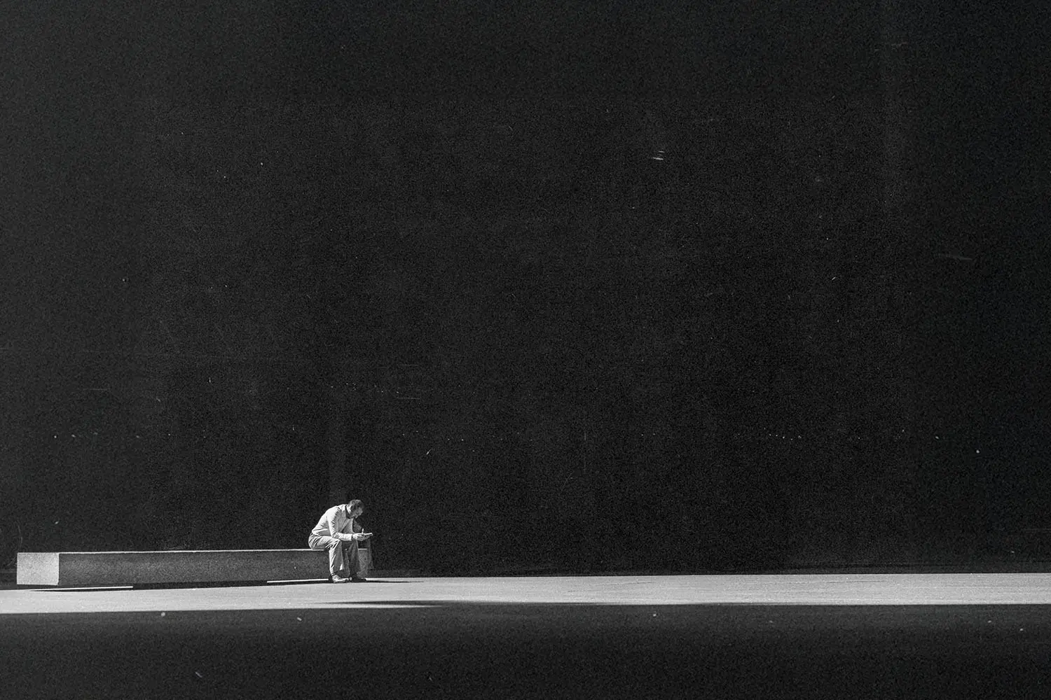 Person sitting alone on a long bench in a vast, dimly lit space, leaning forward with their head down