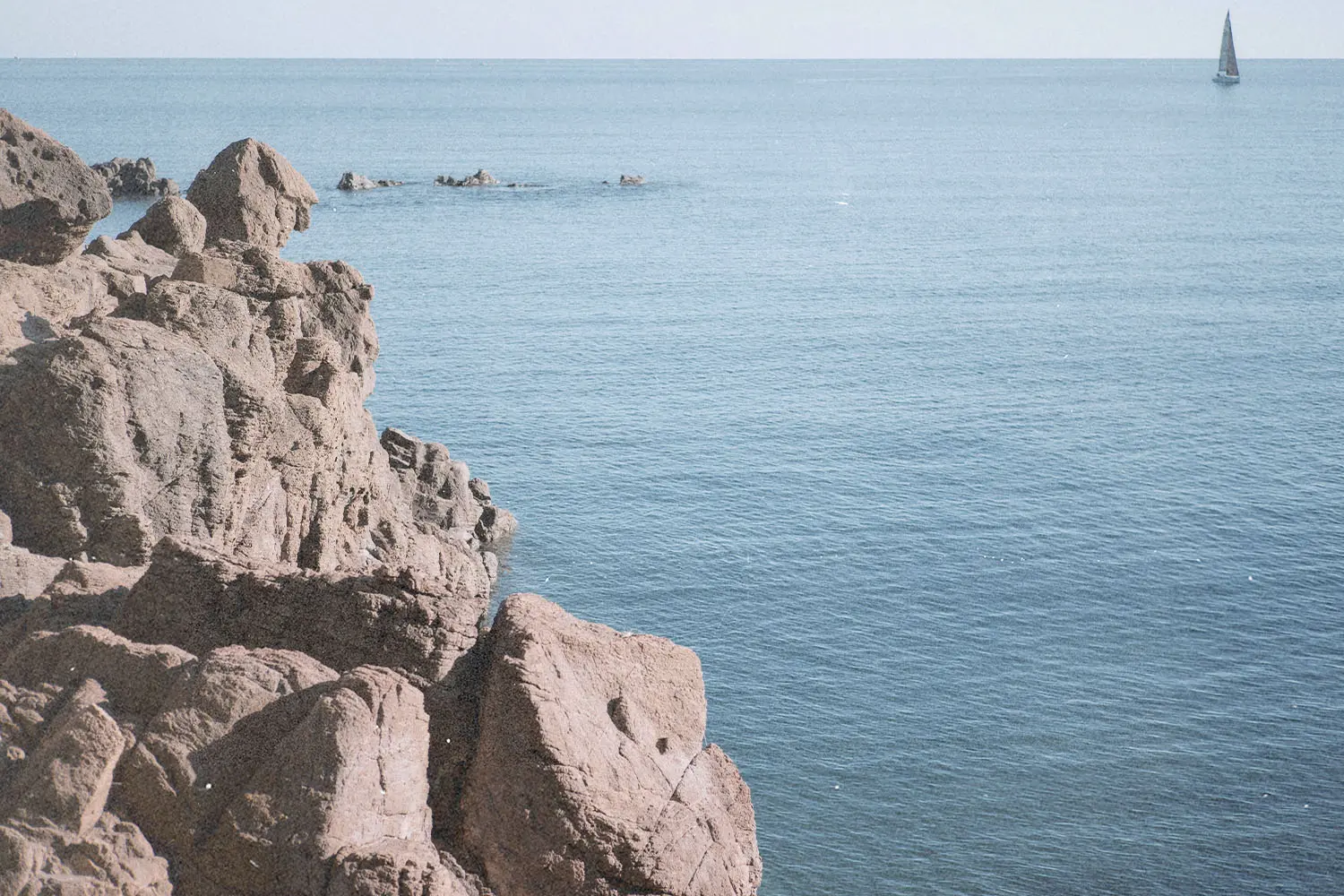 Rocky shoreline overlooking a calm blue sea with a distant sailboat on the horizon
