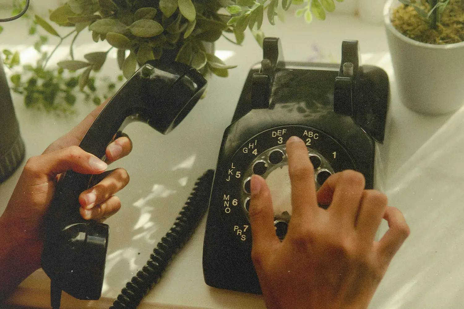 Hands operate a vintage black rotary telephone near potted plants on a bright windowsill