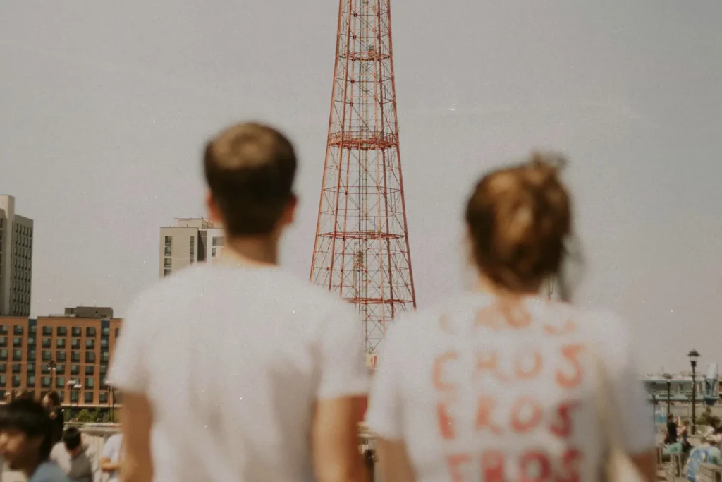 Two people stand outdoors looking toward a tall red metal tower, with buildings and a crowd in the background