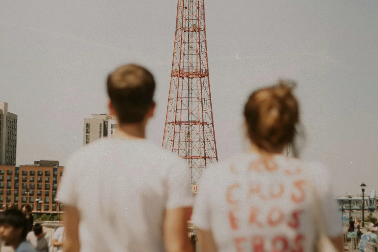 Two people stand outdoors looking toward a tall red metal tower, with buildings and a crowd in the background