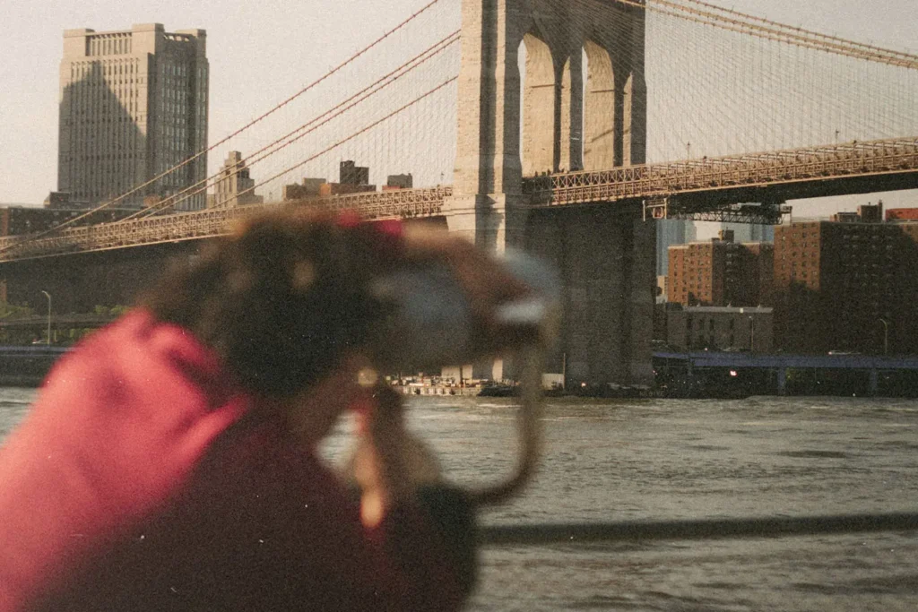 A person looks through a public viewing telescope toward a large suspension bridge crossing over the water