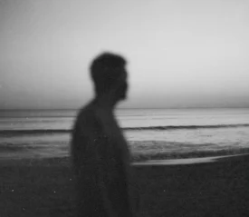 A silhouetted person stands near the shoreline at dusk, with calm waves and a fading horizon behind them