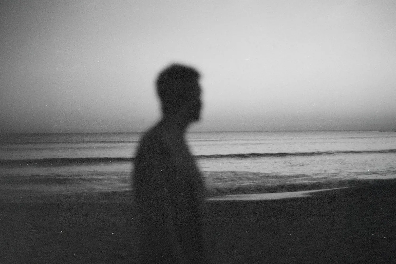 A silhouetted person stands near the shoreline at dusk, with calm waves and a fading horizon behind them