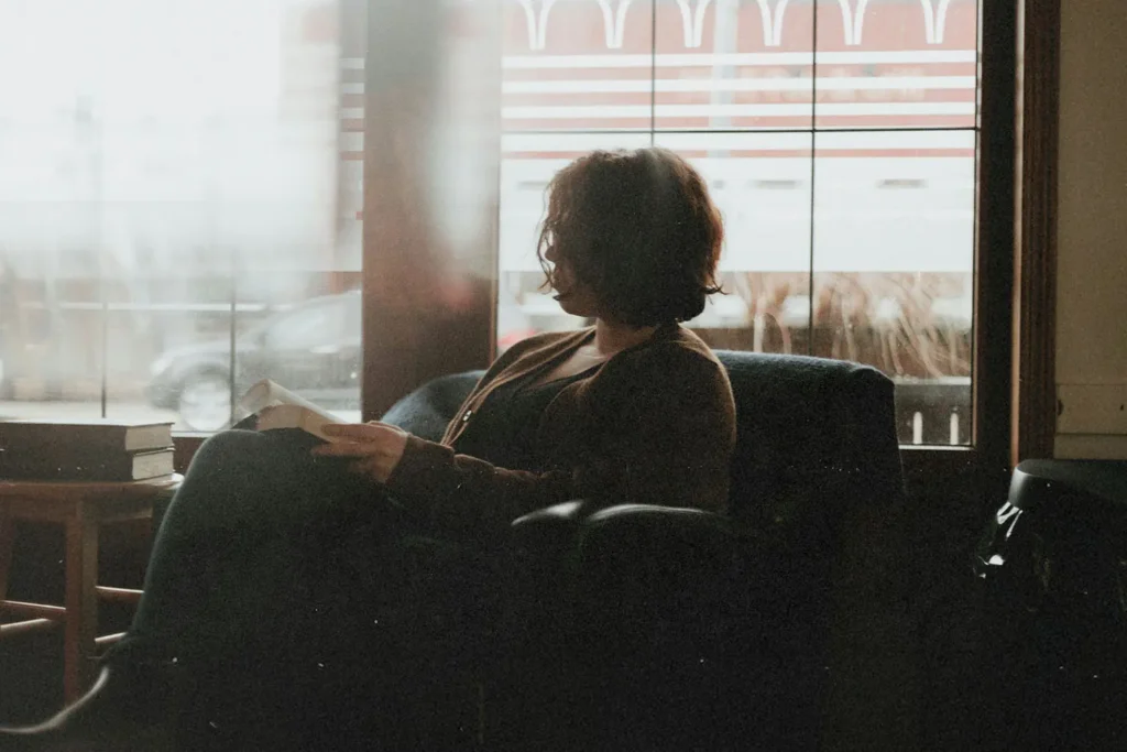 A person sits in a cozy chair near a large window, reading a book as soft daylight filters into the room